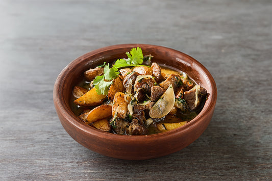 Traditional Georgian Ojakhuri served in a rustic clay bowl, featuring roasted pork, potatoes, and onions garnished with fresh cilantro on a wooden tabletop.